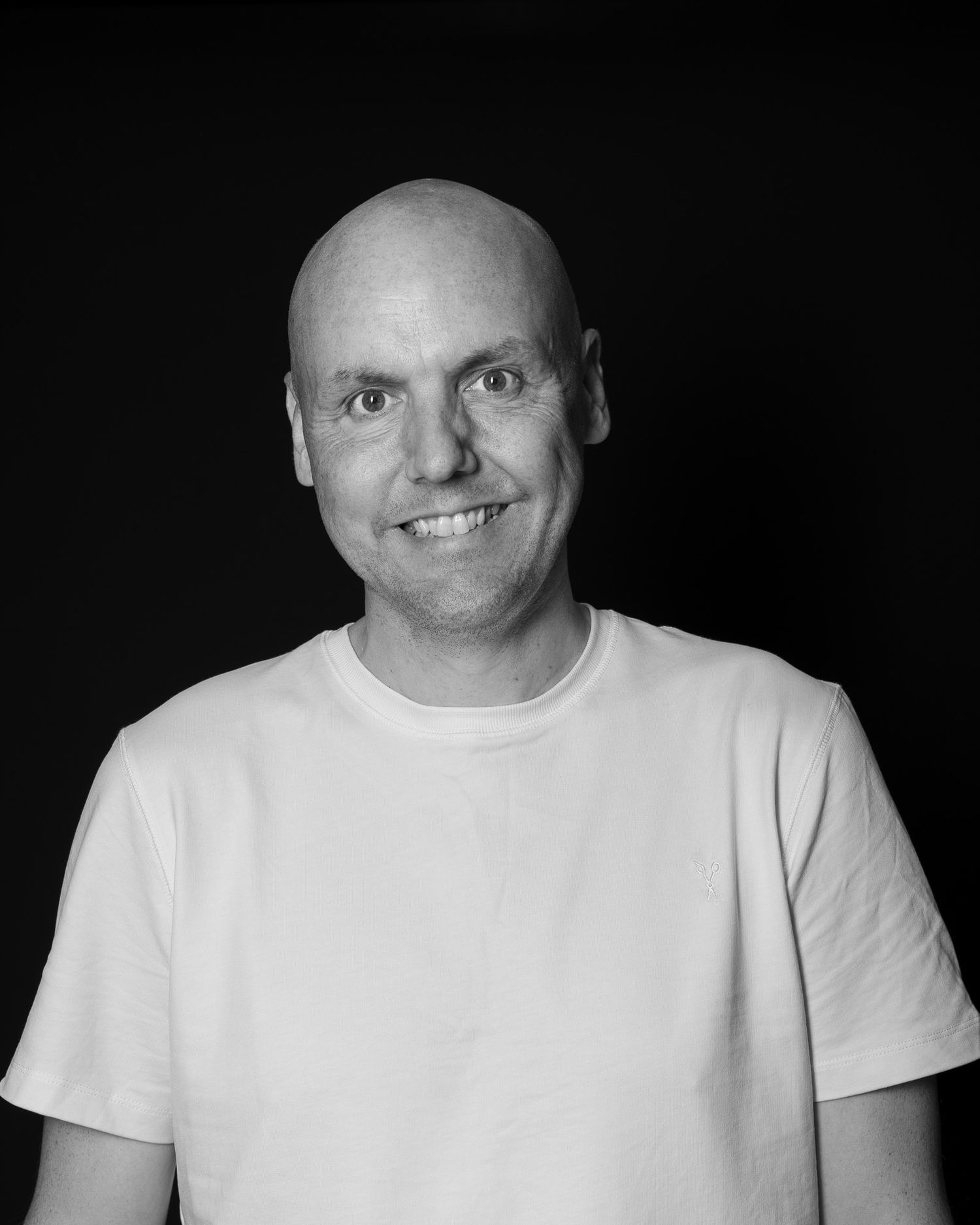 Bald man smiling warmly in a white t-shirt against a dark background, black and white portrait photo.