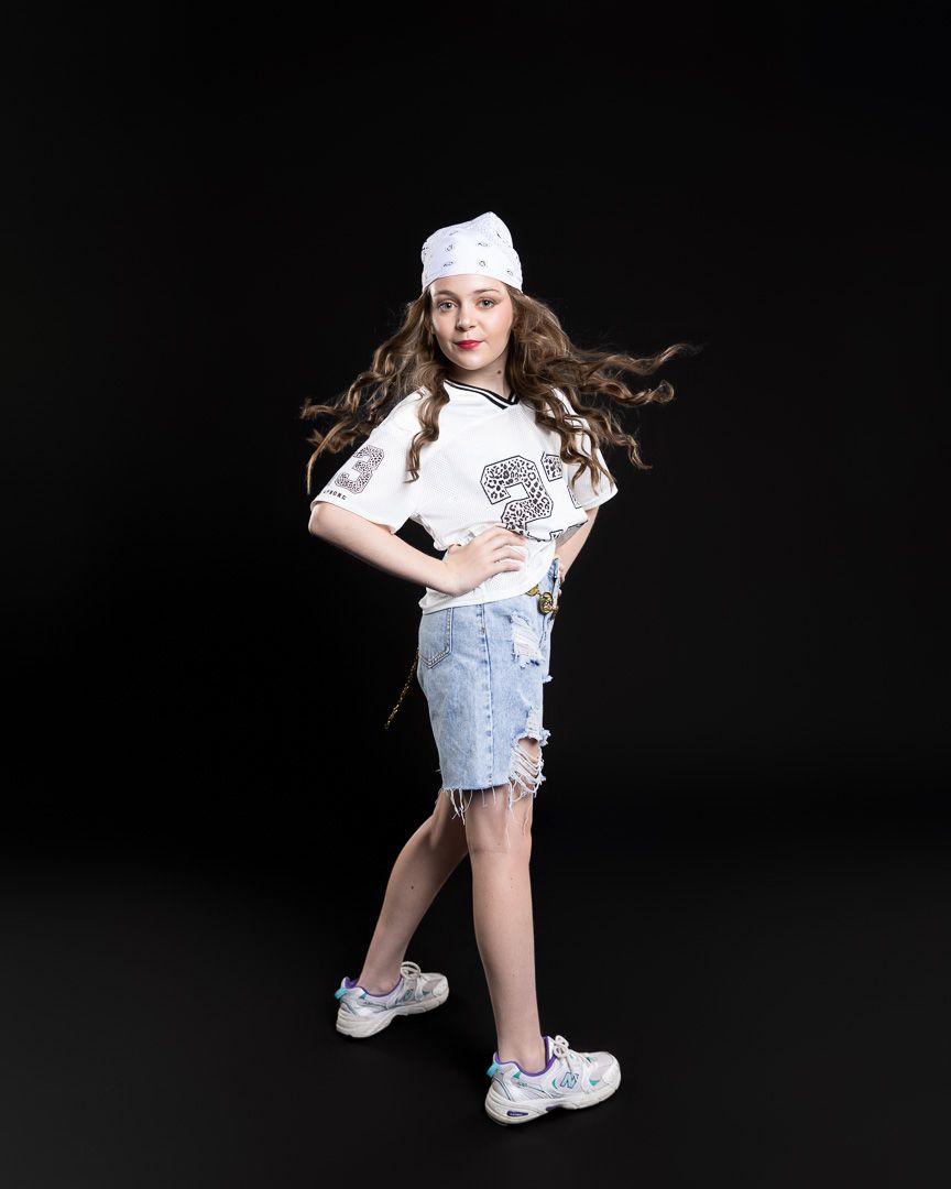 Young girl posing in white graphic tee, denim distressed shorts, white sneakers and headband on dark background.
