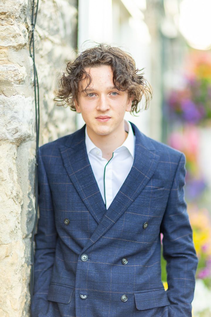 A person wearing a navy blue double-breasted suit jacket and white shirt poses against a stone wall backdrop.