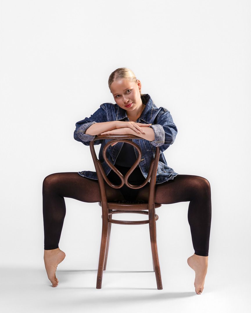 Barefoot dancer in denim jacket and black tights poses straddling a wooden chair against white background.