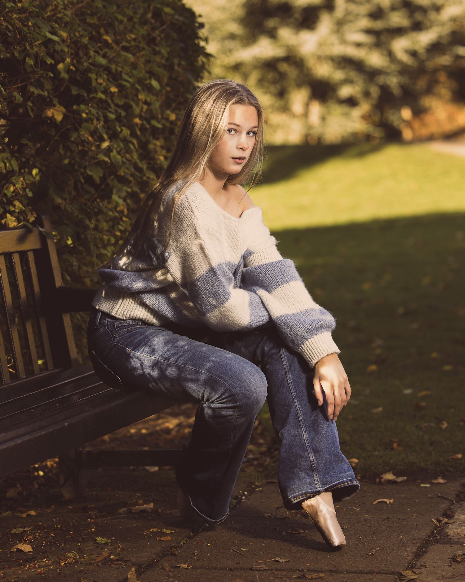 A person in casual attire sits on a wooden bench in a sunlit autumn garden setting.