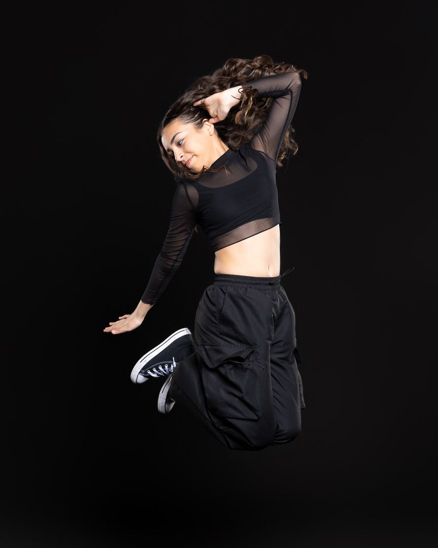 A dancer leaps against a black background, wearing a black crop top and pants with sneakers.