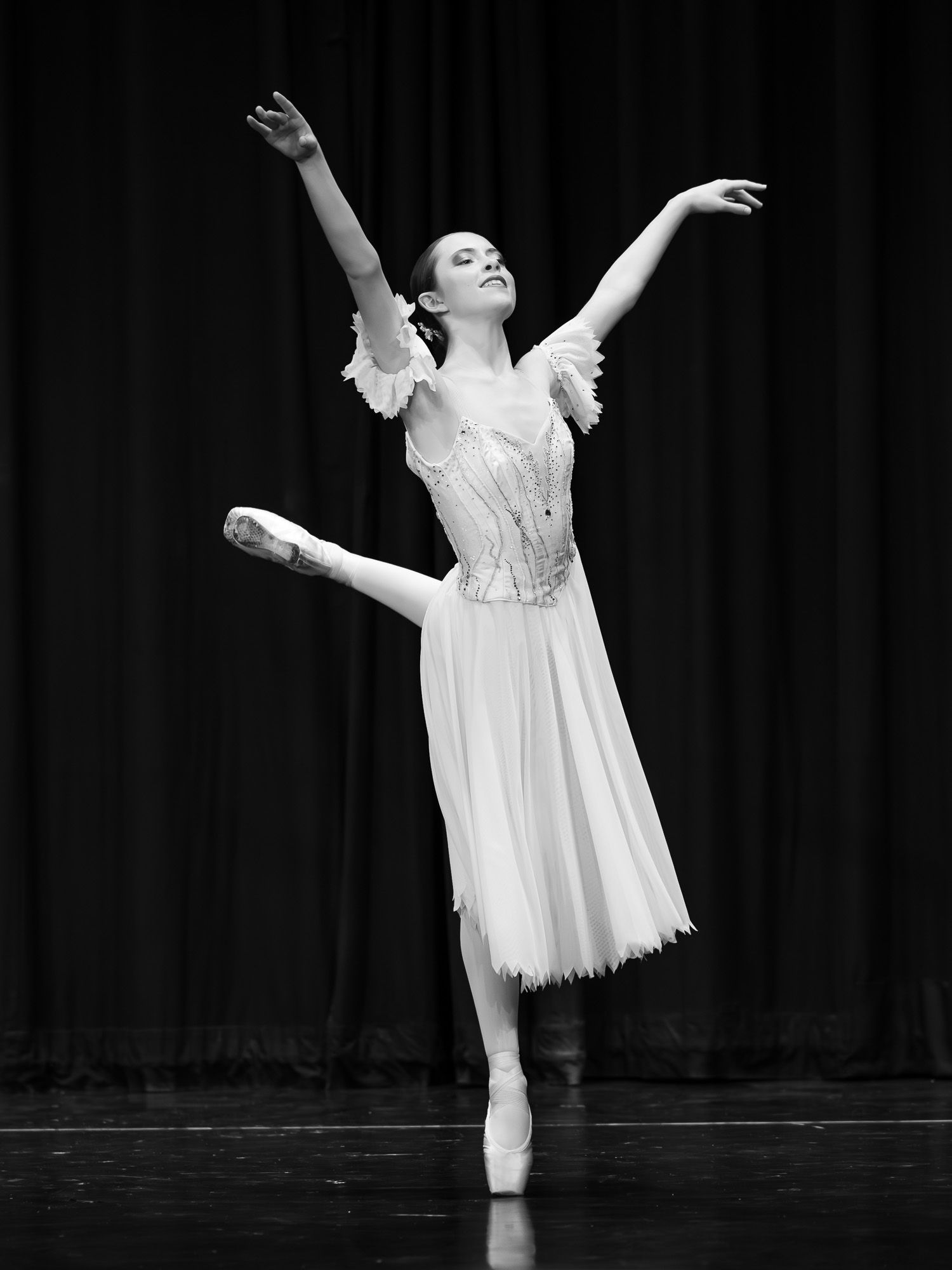 Ballerina in white dress performs elegant arabesque on pointe on stage.