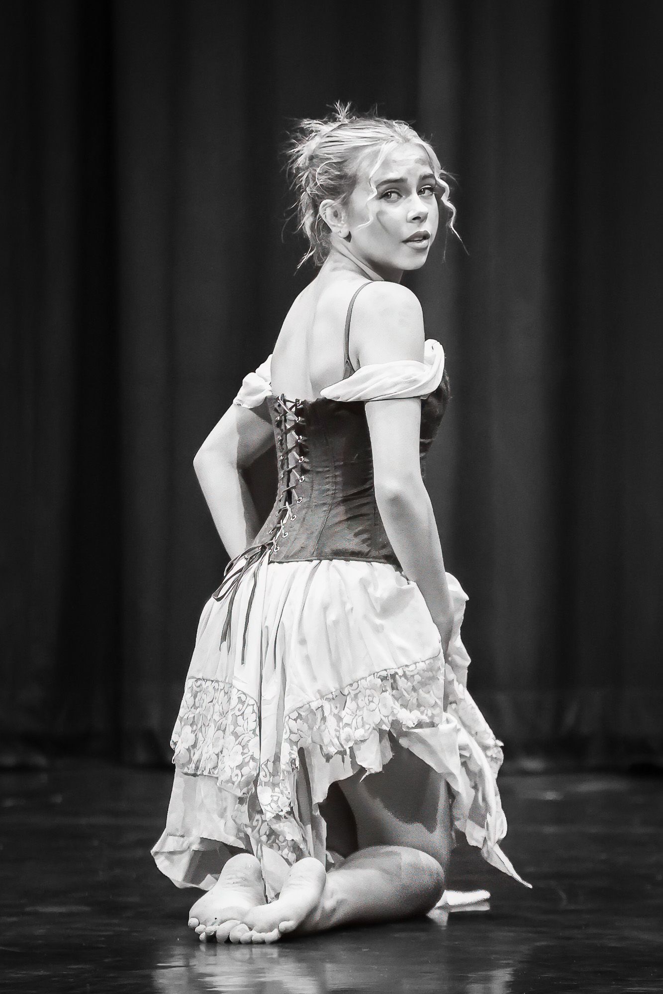 Black and white photo of a female dancer in a corset dress kneeling on stage, looking over her shoulder.