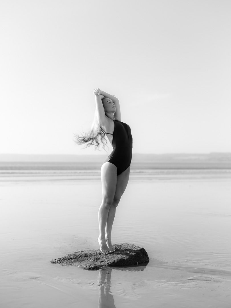 Black and white photo of woman in swimsuit posing gracefully on rock at beach shoreline.