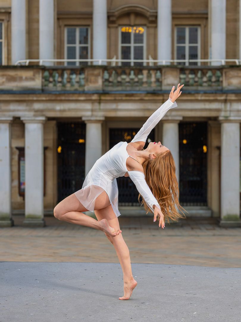 A dancer in a white dress performs an elegant arabesque pose outdoors before a classical stone building.