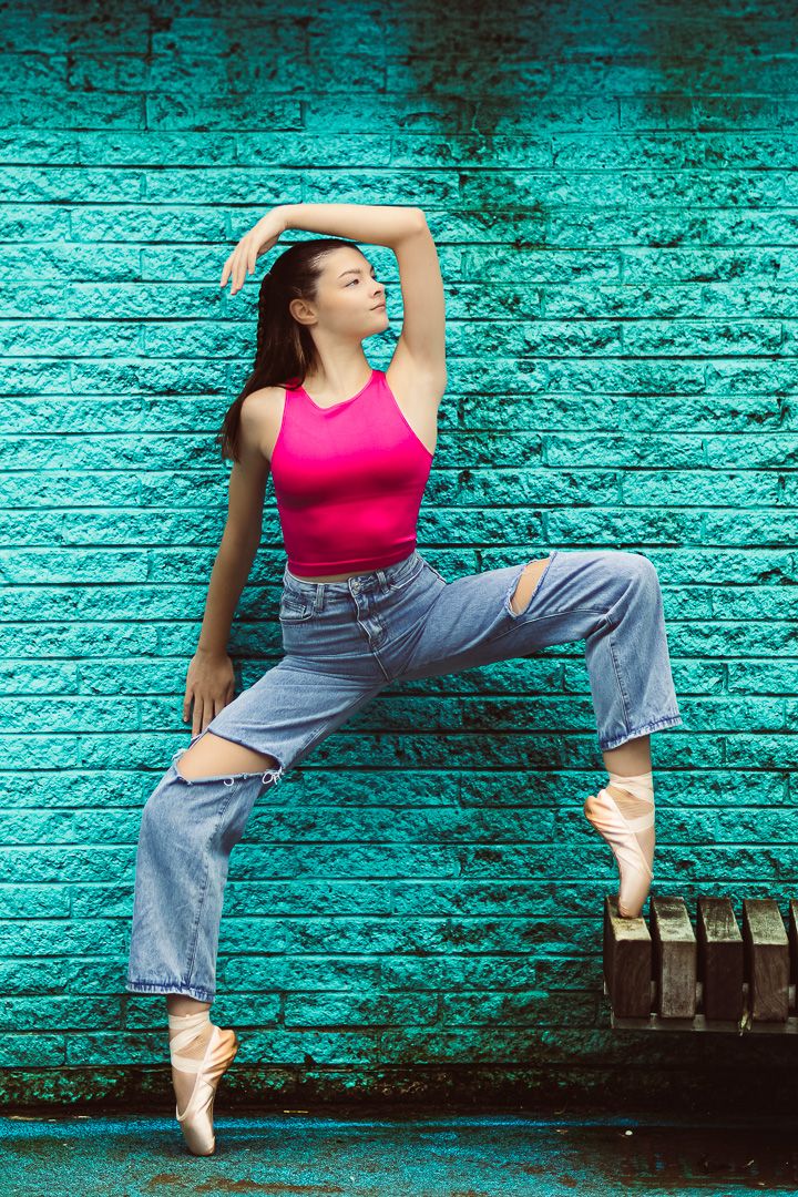 A ballet dancer in pink top and ripped jeans poses en pointe against a turquoise brick wall.