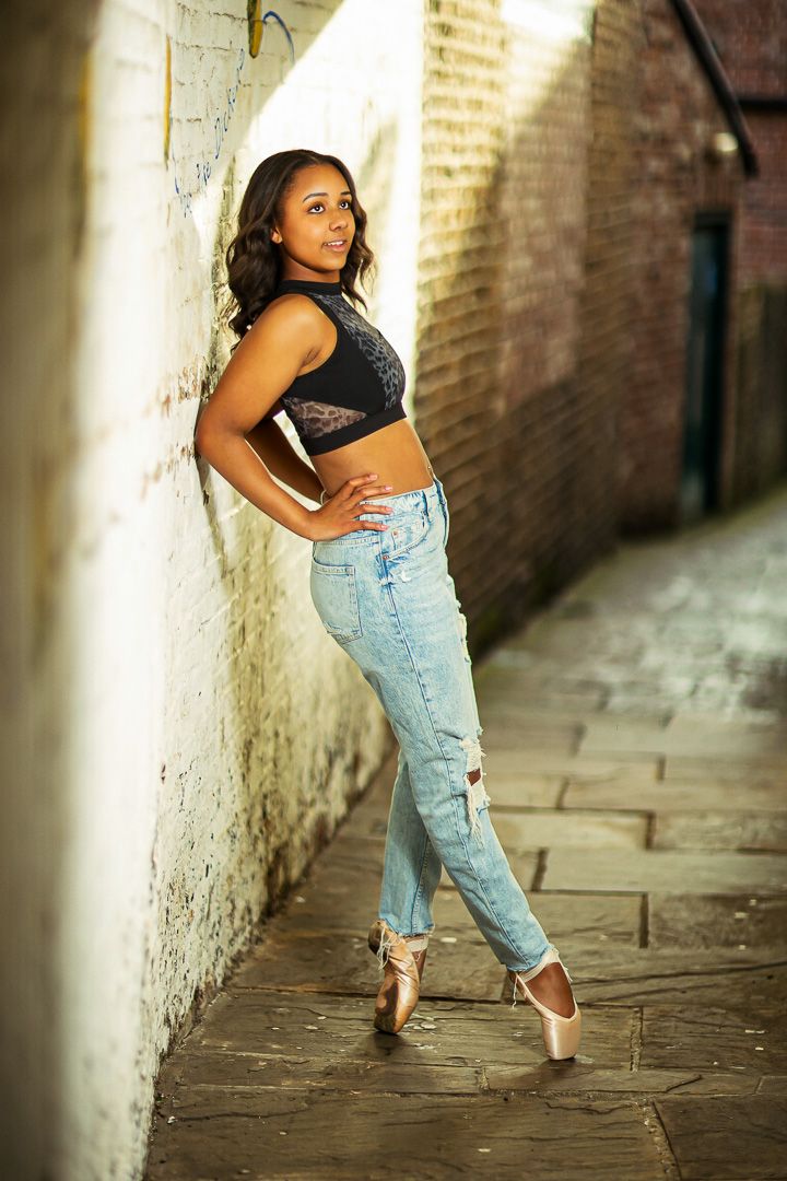 Dancer in pointe shoes performs in narrow alley with dramatic lighting.