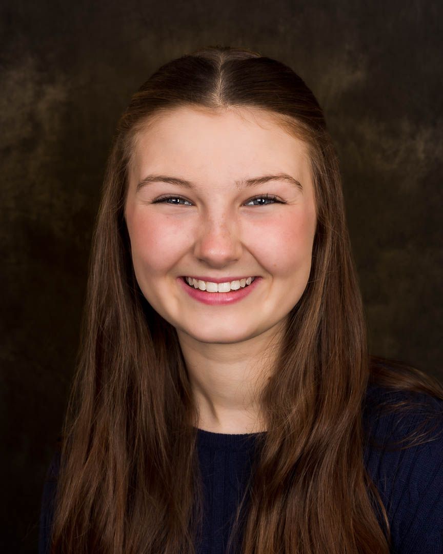 Smiling young woman with long brown hair wearing a navy top, professional headshot portrait.