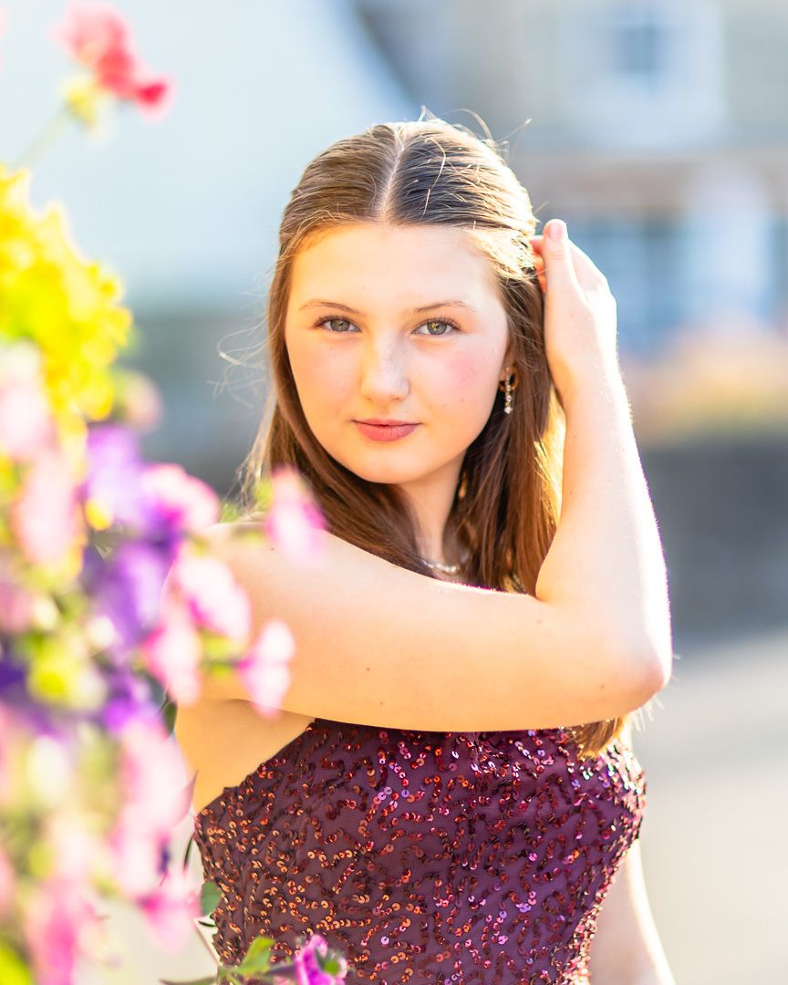 A young woman in a sparkly burgundy dress poses outdoors next to colorful flowers on a sunny day.