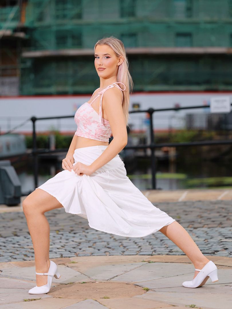 Dancer in white dress performs ballet poses on cobblestone street.