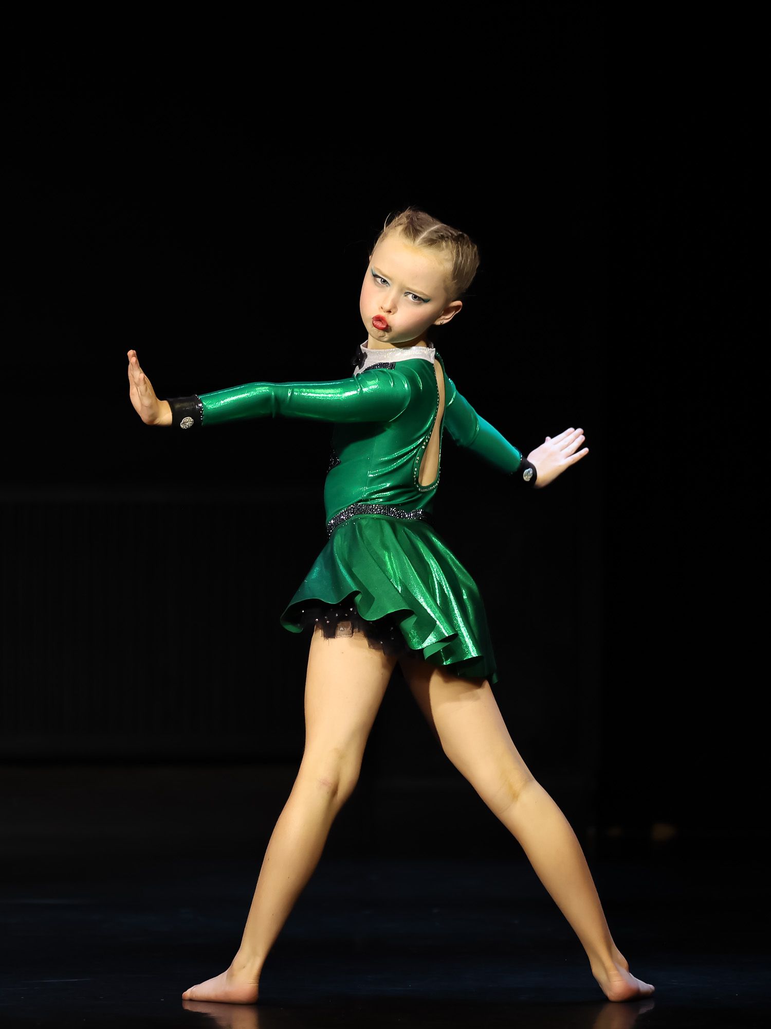 Young girl performing on stage in a green metallic dance costume, striking a dramatic pose under stage lighting.