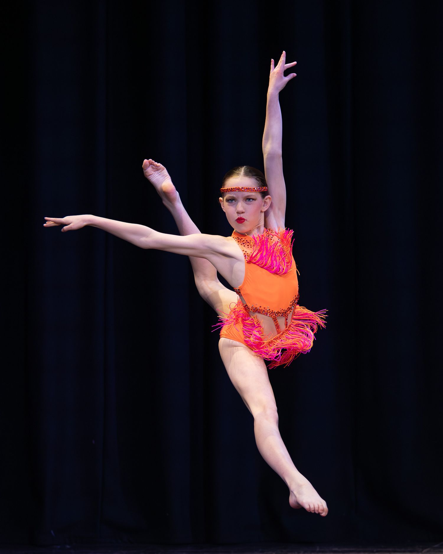 Young dancer leaps gracefully in vibrant orange costume with fringe details under stage lights.
