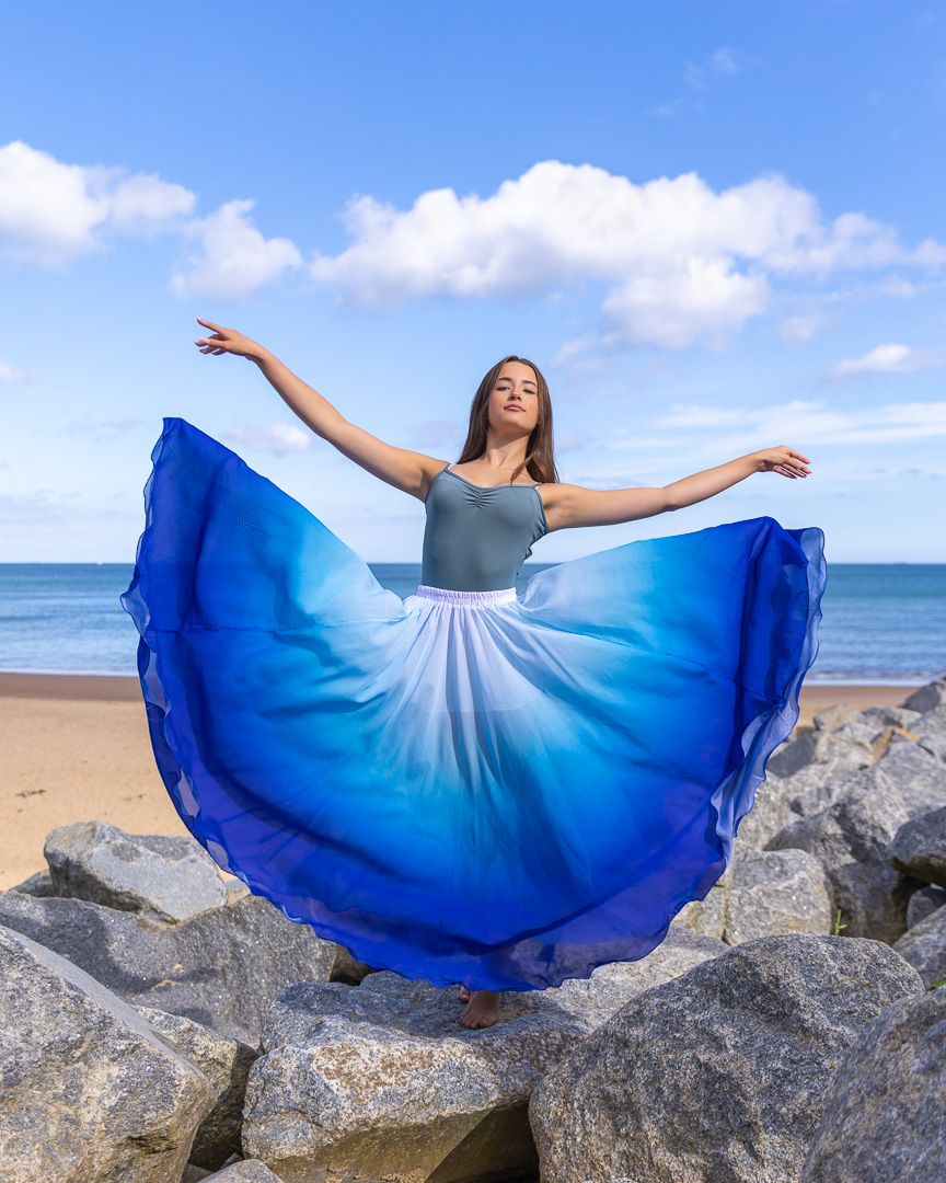 Dancer in blue ombre skirt poses gracefully on rocks at beach, arms outstretched under blue sky.