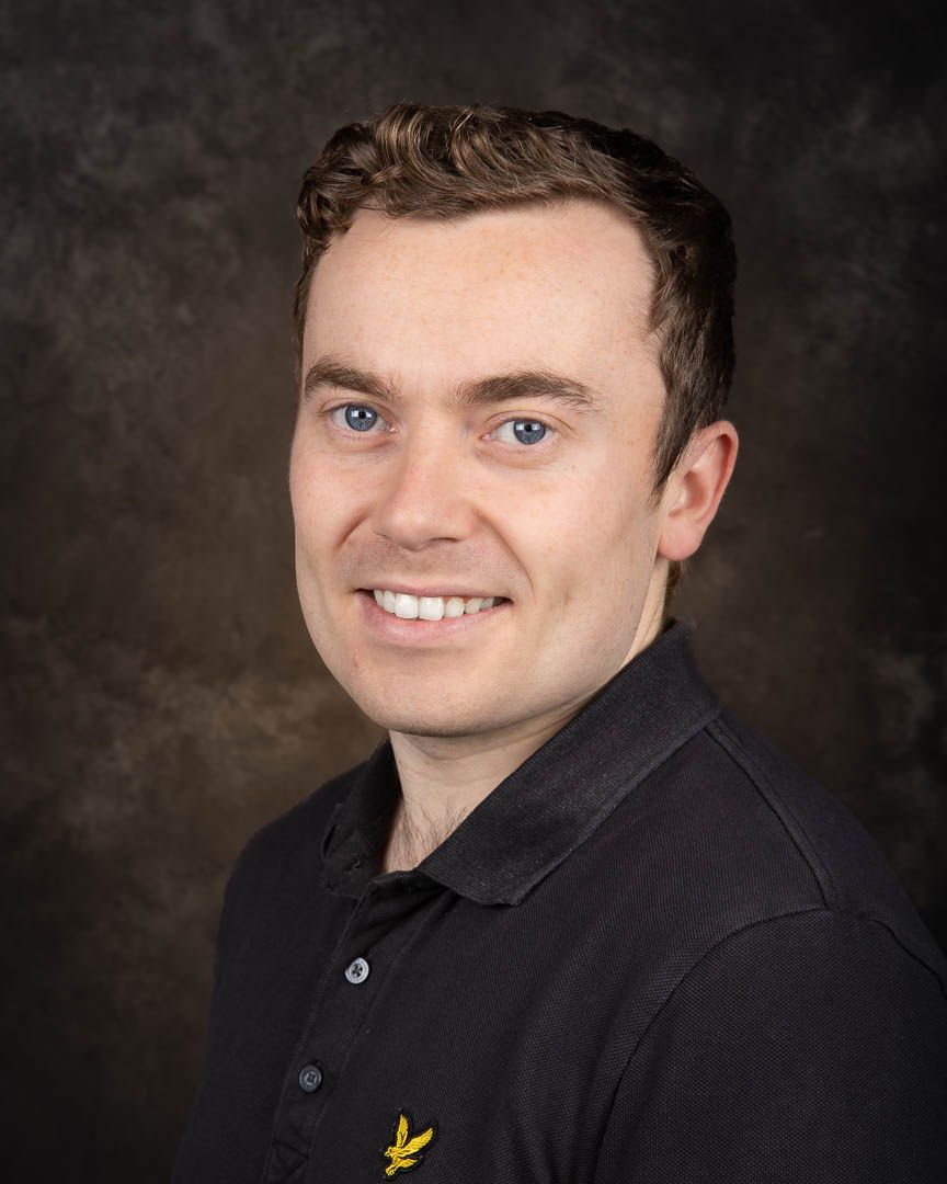 Smiling young man with blue eyes wearing a black polo shirt in a professional headshot.