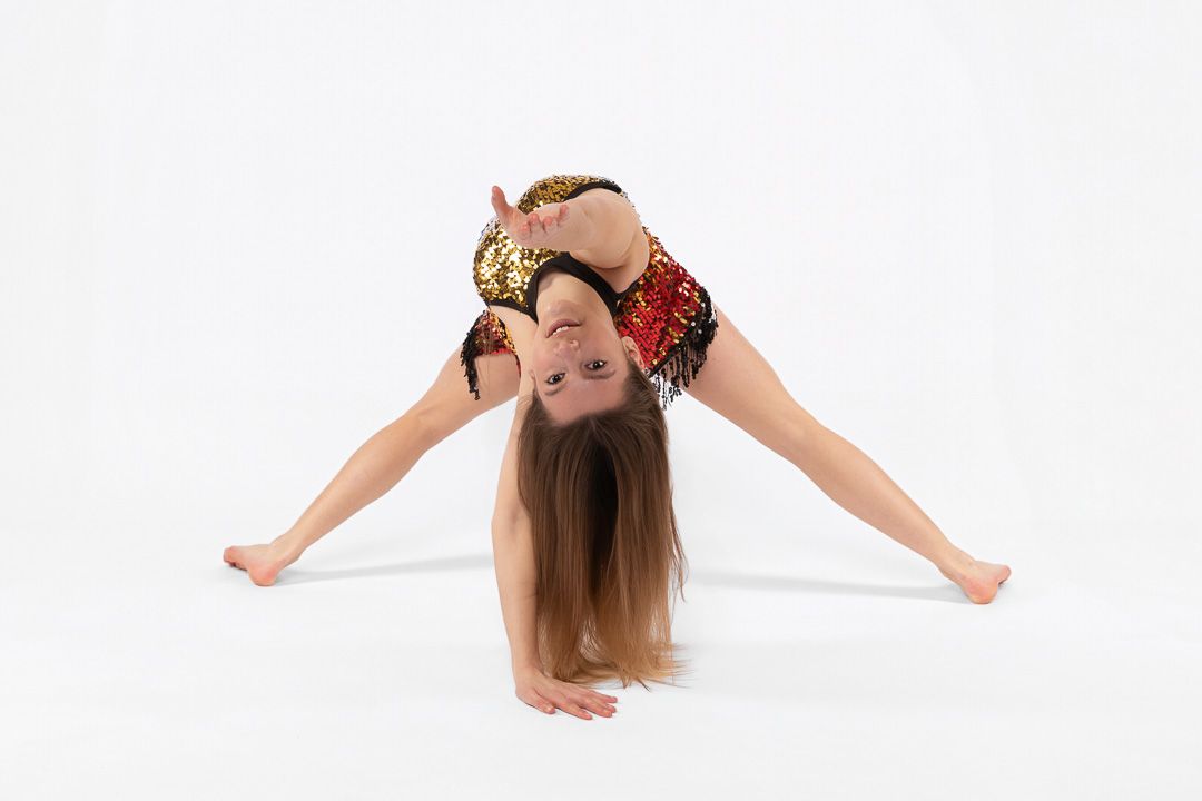 Young gymnast in red and gold leotard performing a backbend split on white background.