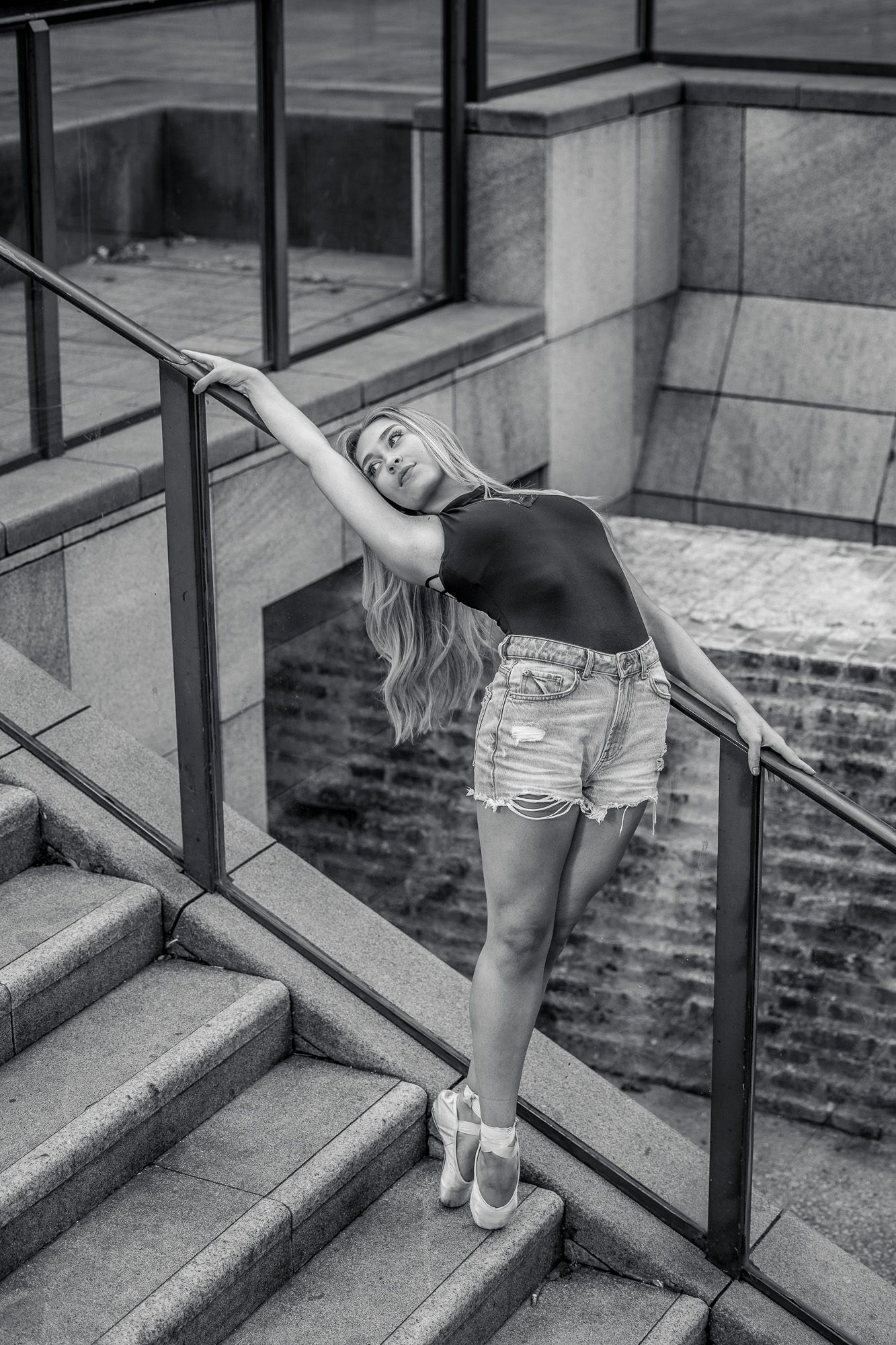 A dancer in ballet shoes and denim shorts strikes a graceful pose while leaning back on an outdoor staircase railing.
