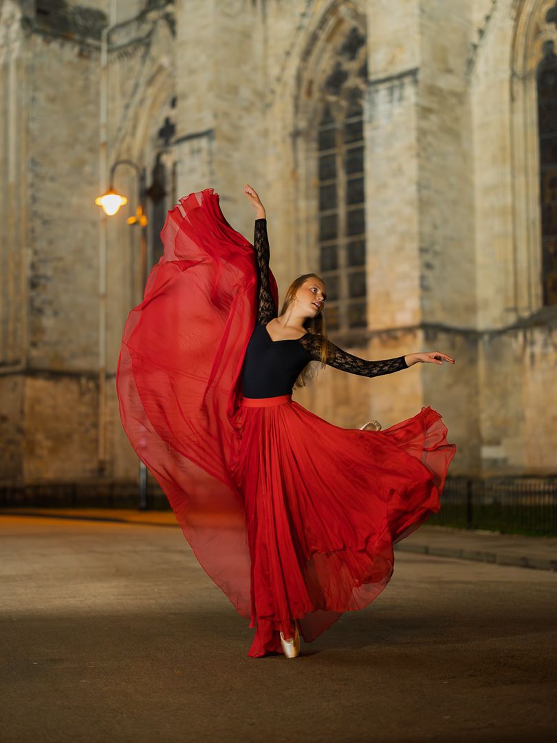 A flowing red dress twirls gracefully against the backdrop of a historic stone cathedral at dusk.