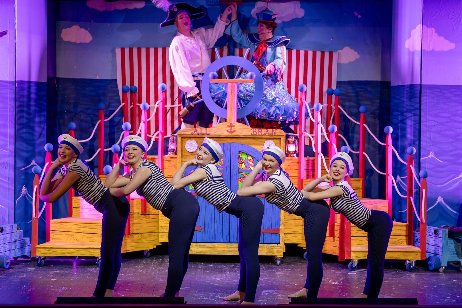 Sailors in striped costumes perform a high-kick dance routine on a colorful nautical-themed stage set.