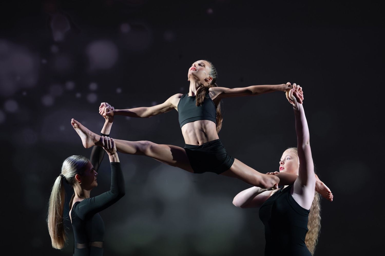 Three female dancers perform an acrobatic lift routine against a dark background, showcasing flexibility and strength.