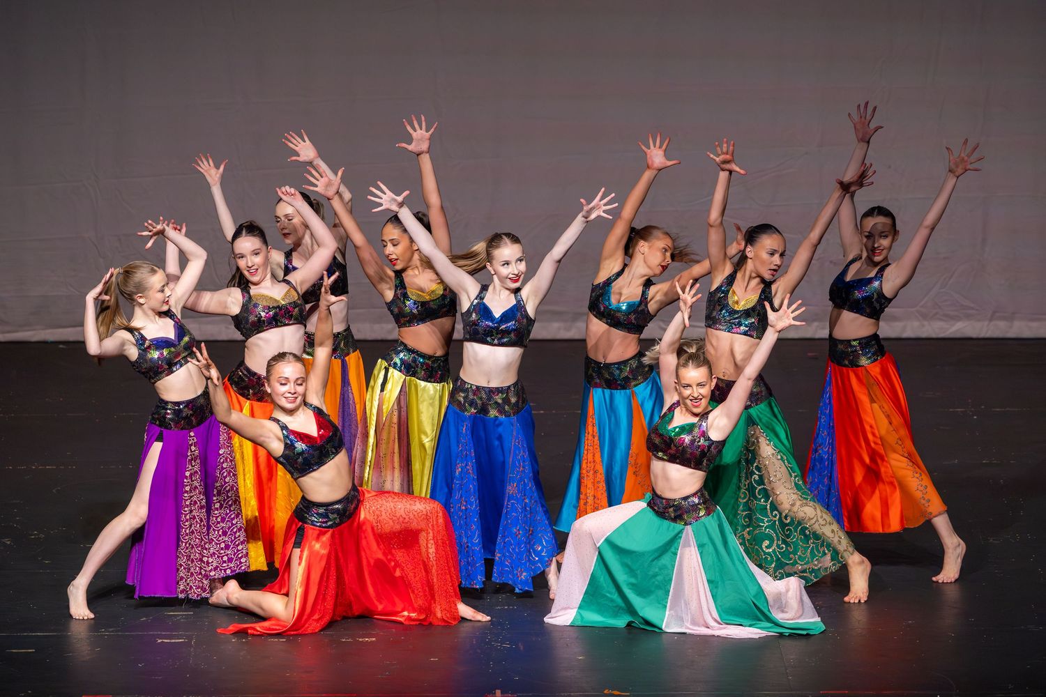 Young girls perform a colorful belly dance routine on stage in vibrant costumes with raised arms.