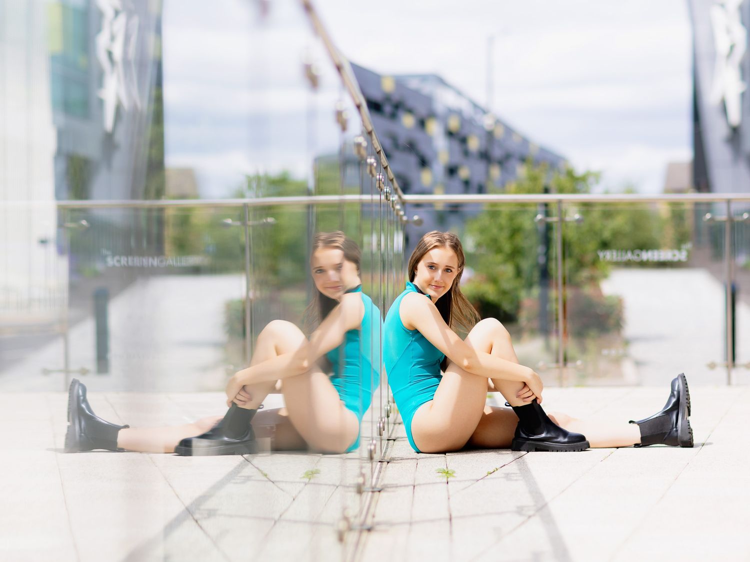 Flexible dancer in teal leotard and black boots sits in splits pose reflected in glass wall outdoors.