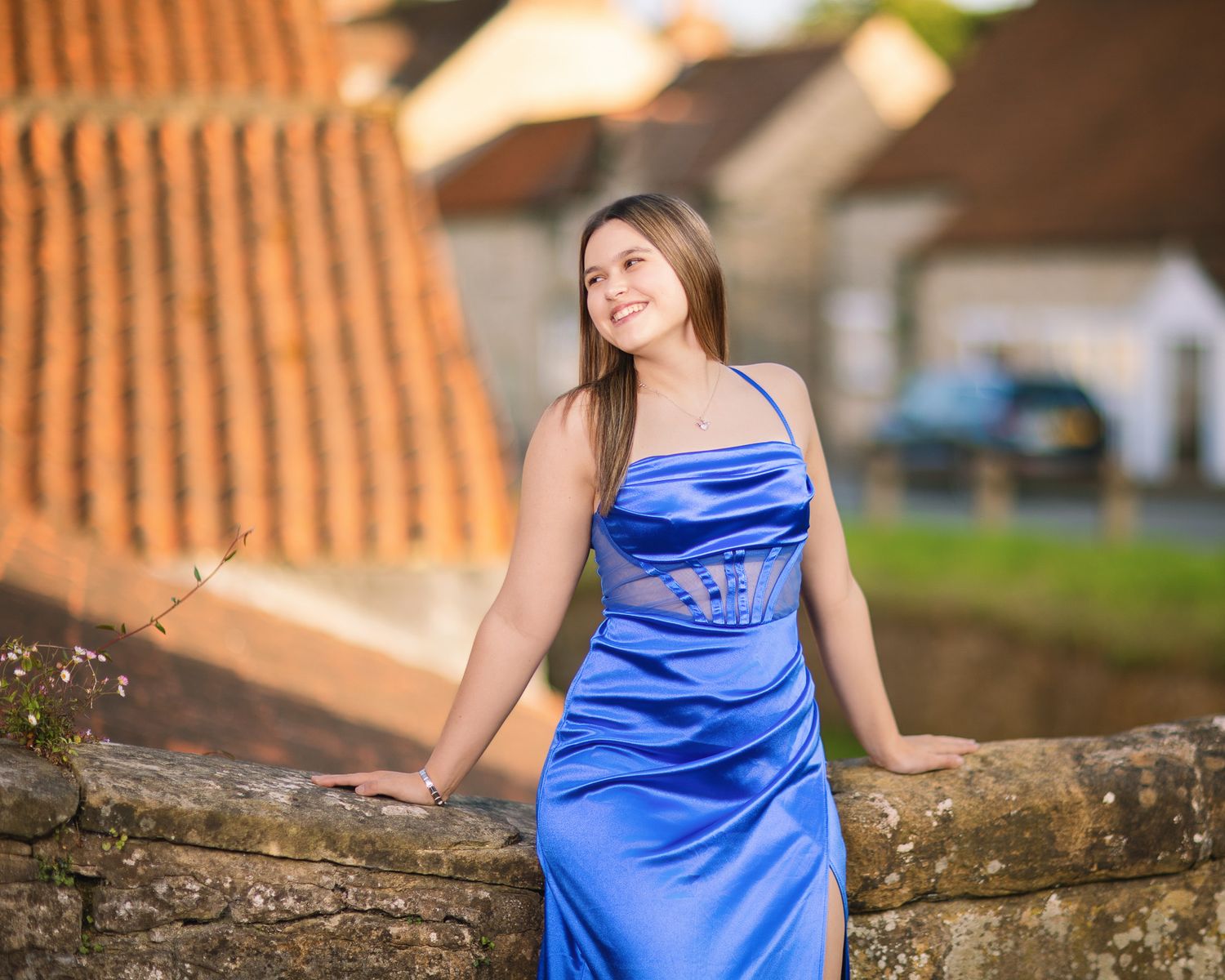 A woman in a royal blue satin dress poses on a rustic stone wall in an outdoor setting.