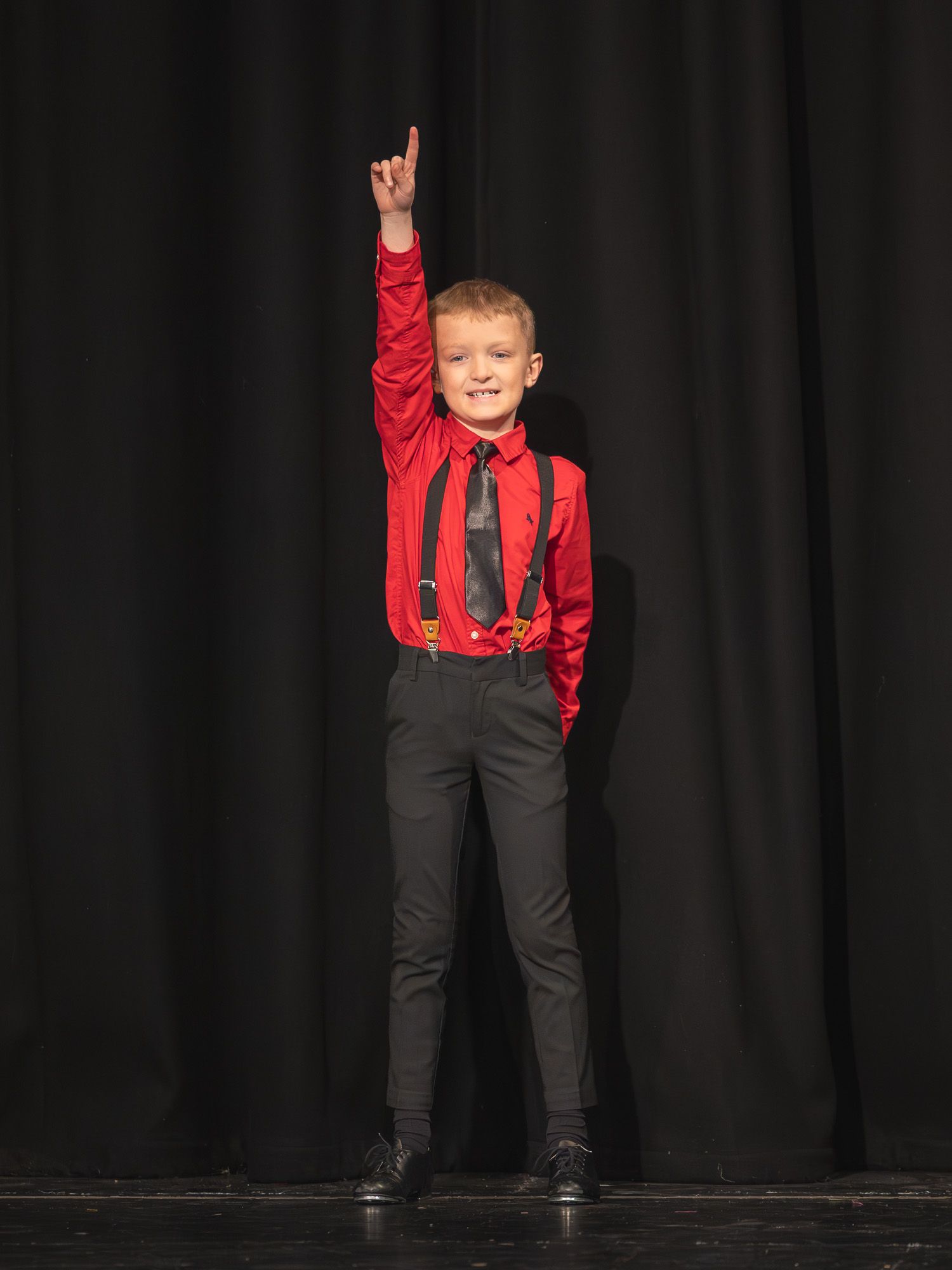 Young boy in red shirt, suspenders, and tie poses on stage with one finger pointed up.