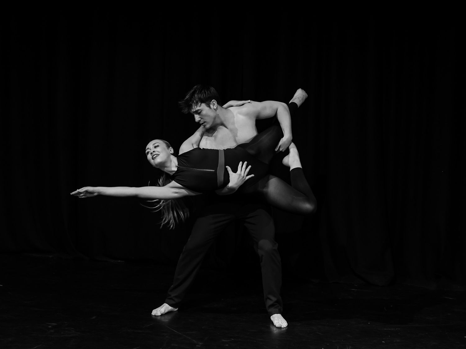 Two dancers perform an elegant contemporary ballet lift pose on a dark stage in a black and white photo.