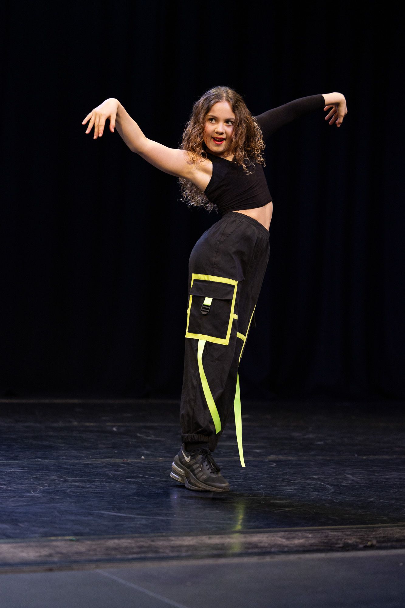 Young dancer performs hip-hop routine on stage in black crop top and neon yellow cargo pants.