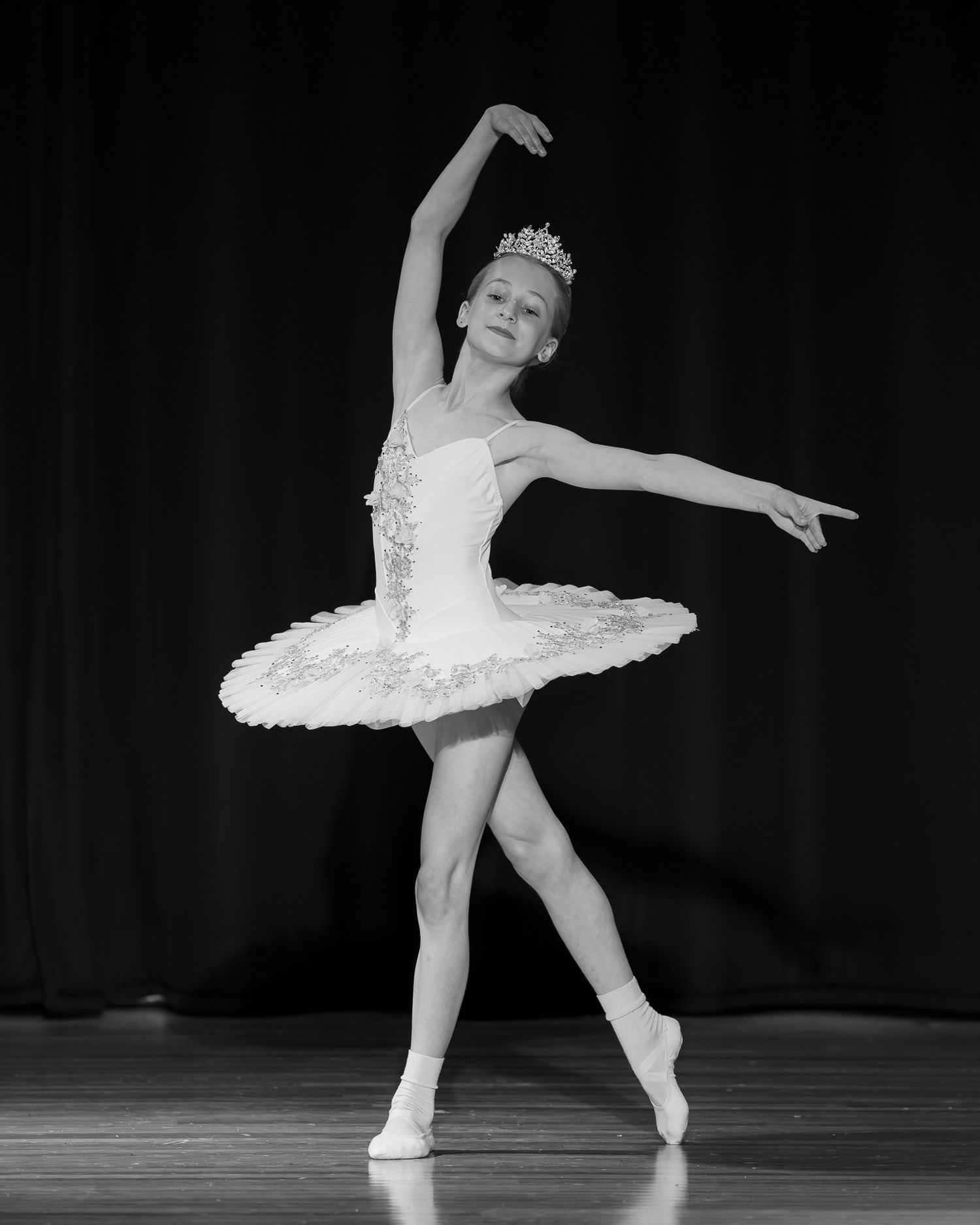 Young ballerina in white tutu and tiara performs graceful arabesque pose on stage in black and white photo.