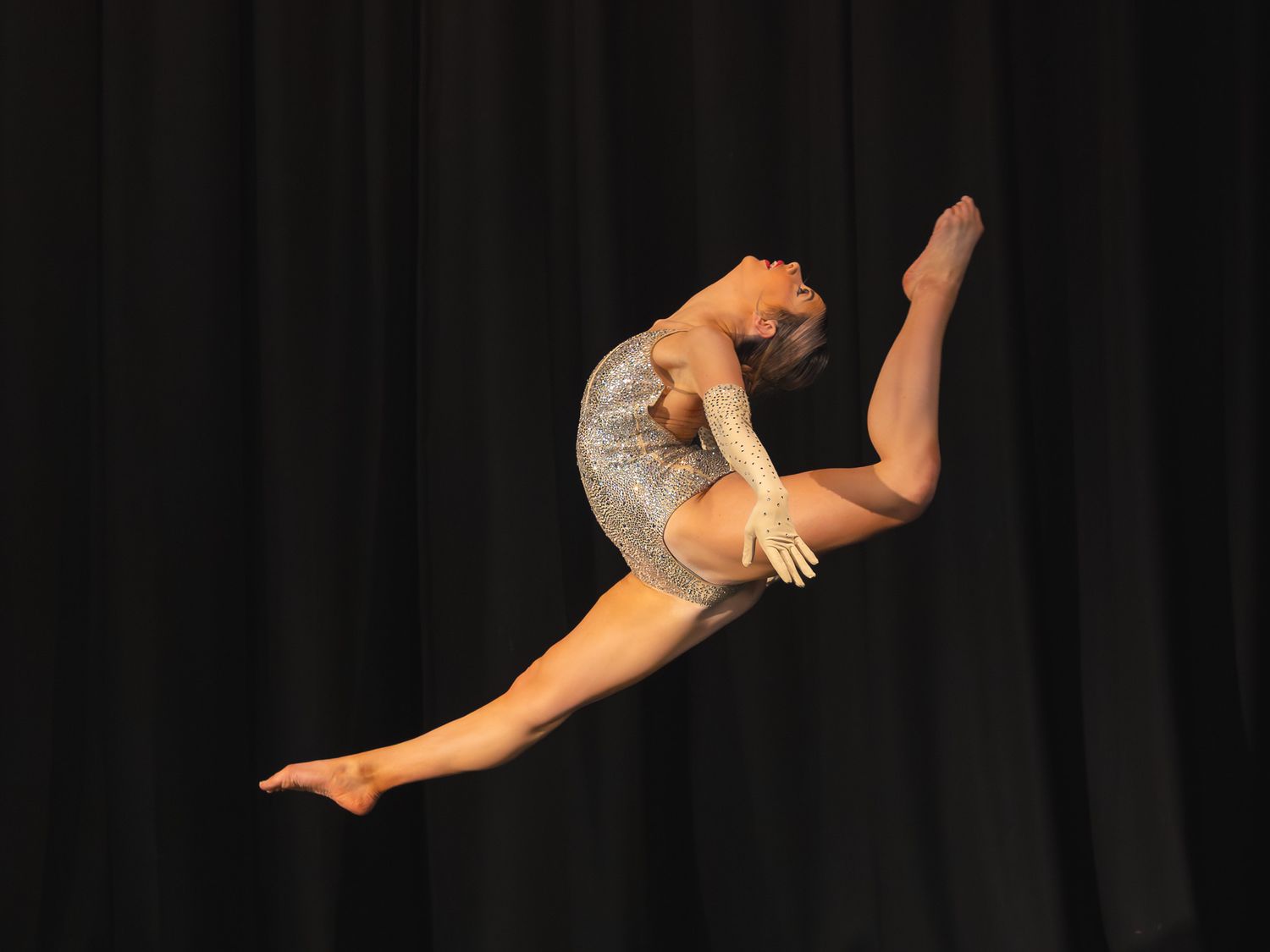 Gymnast performing a mid-air split leap in a silver leotard against a dark stage backdrop.