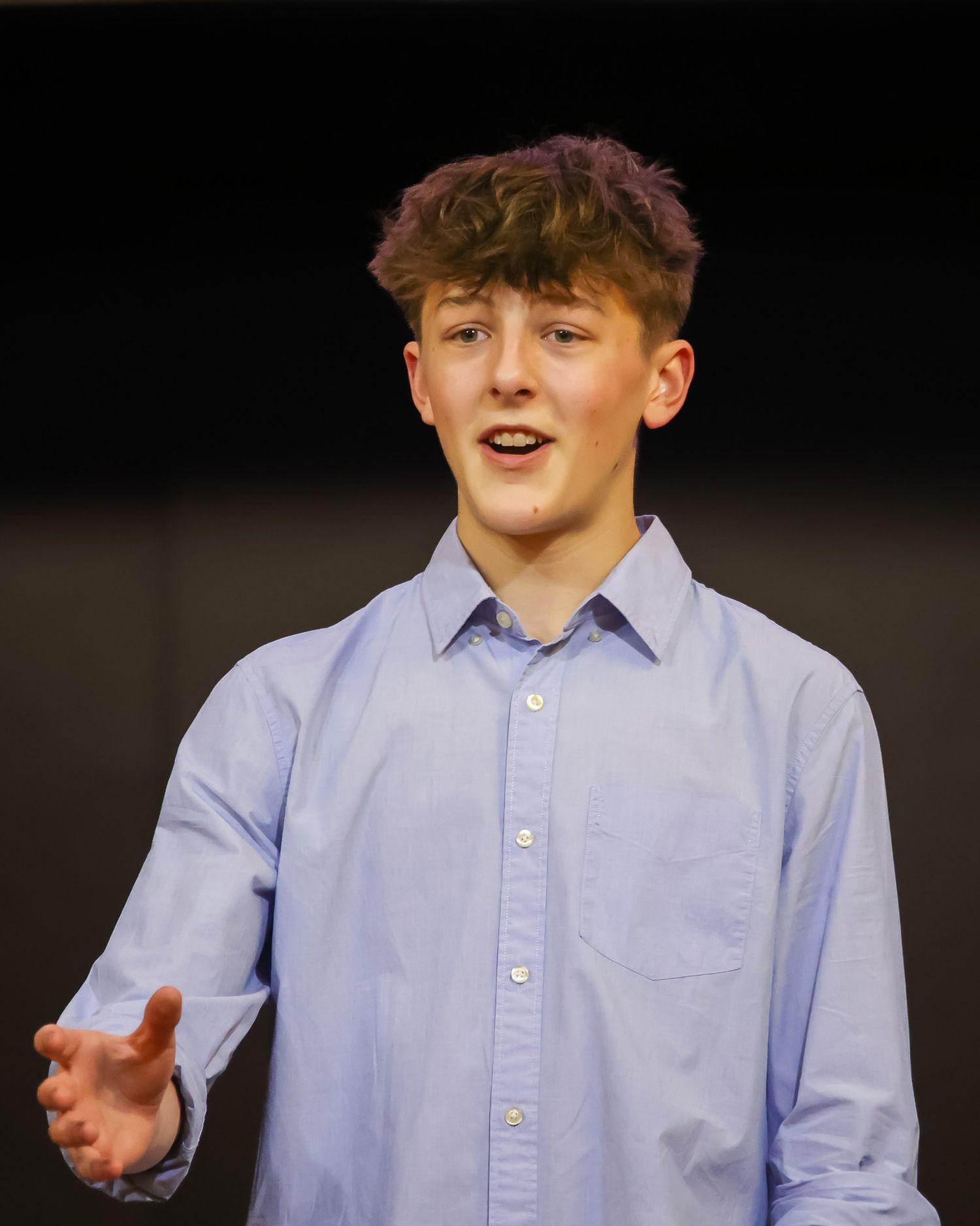 Teenage boy in light blue button-up shirt gesturing while performing on a dark stage.