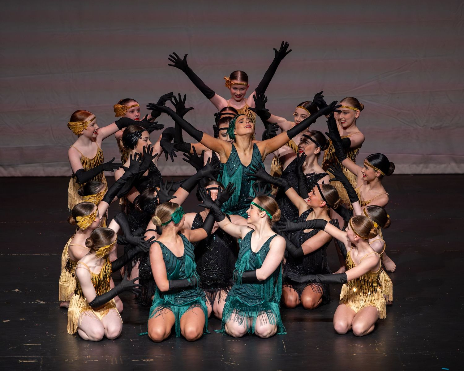 Dancers in gold and green flapper costumes perform a dramatic group pose on stage during a jazz dance recital.