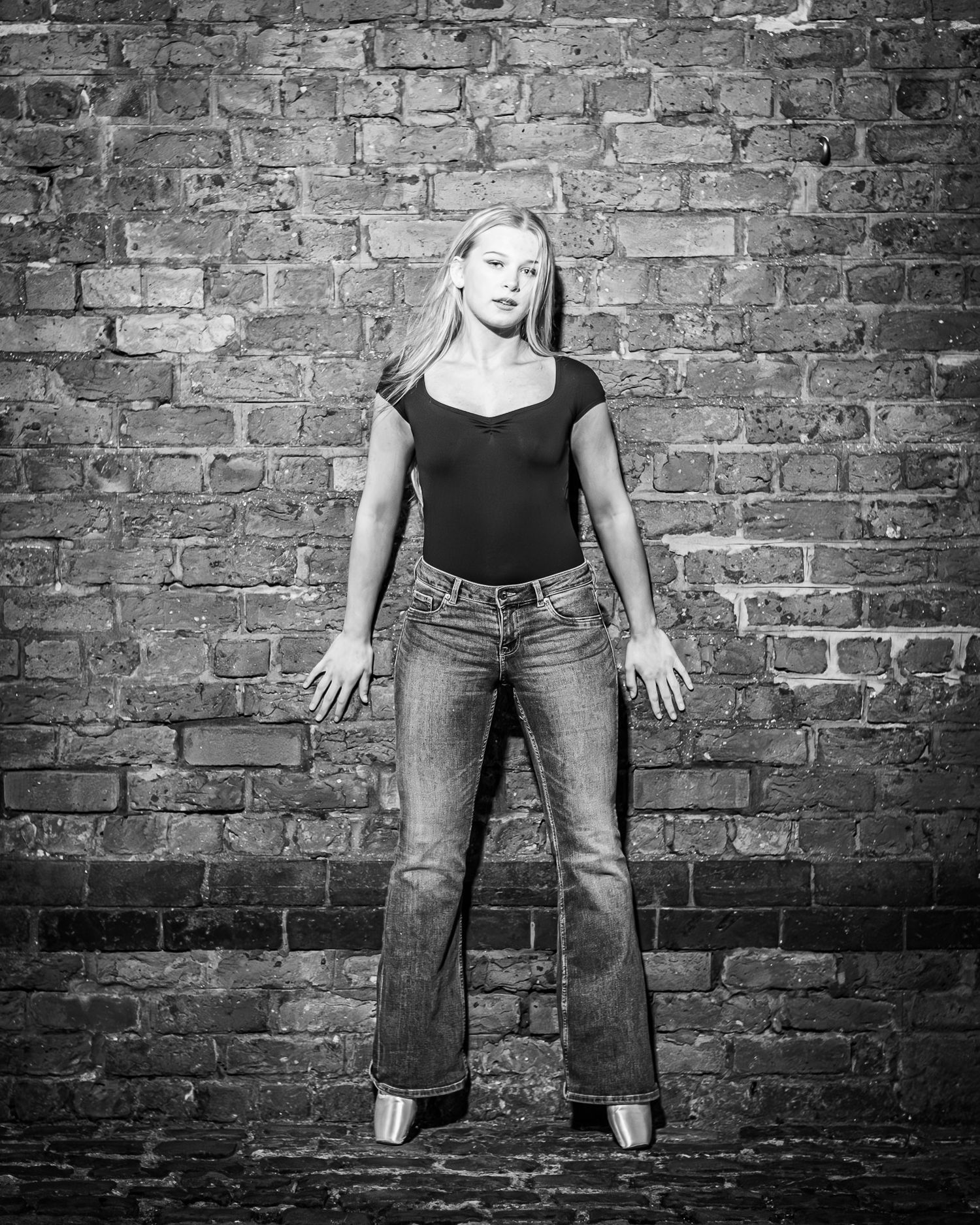 Black and white photo of a young blonde woman posing confidently in jeans and black top against a brick wall.