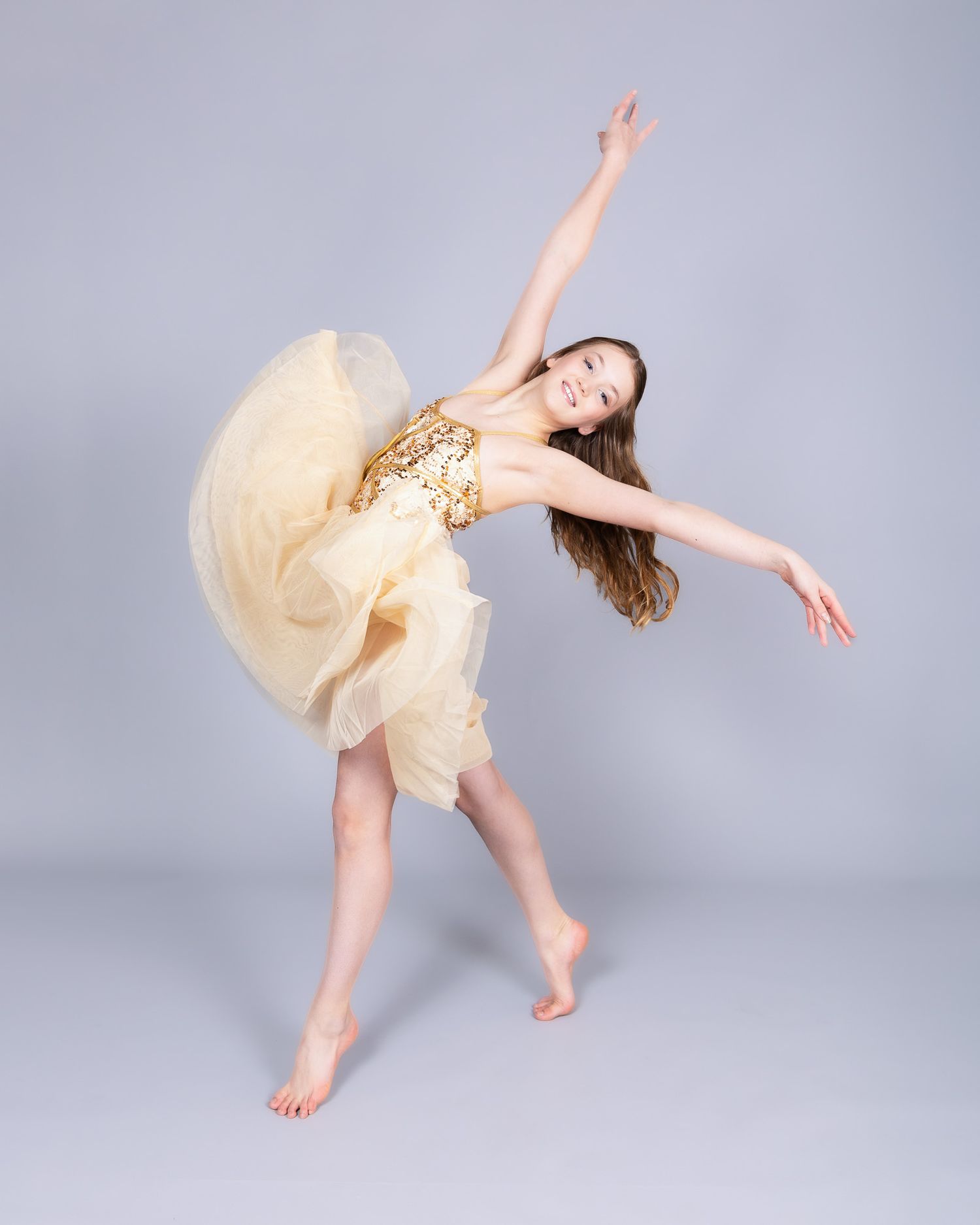 Dancer in flowing gold and cream dress performs graceful barefoot movement against gray studio backdrop.
