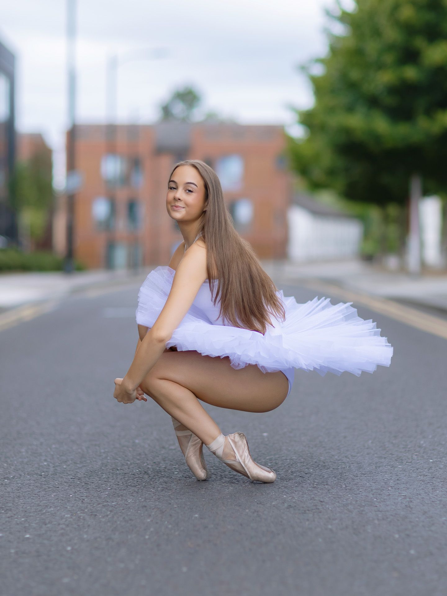A young ballerina in a white tutu and pointe shoes poses gracefully on an urban street.