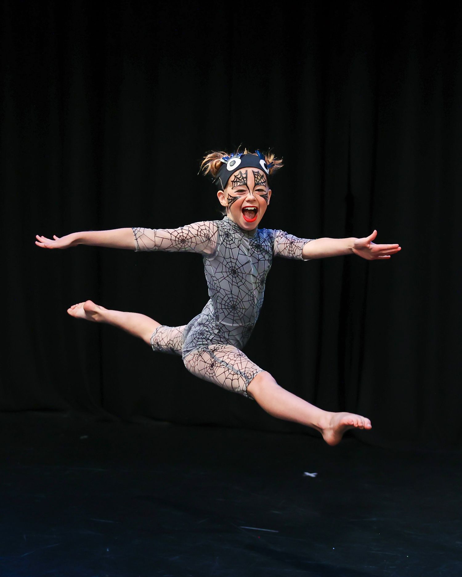 Young dancer leaps mid-air in spider web costume with face paint on dark stage.