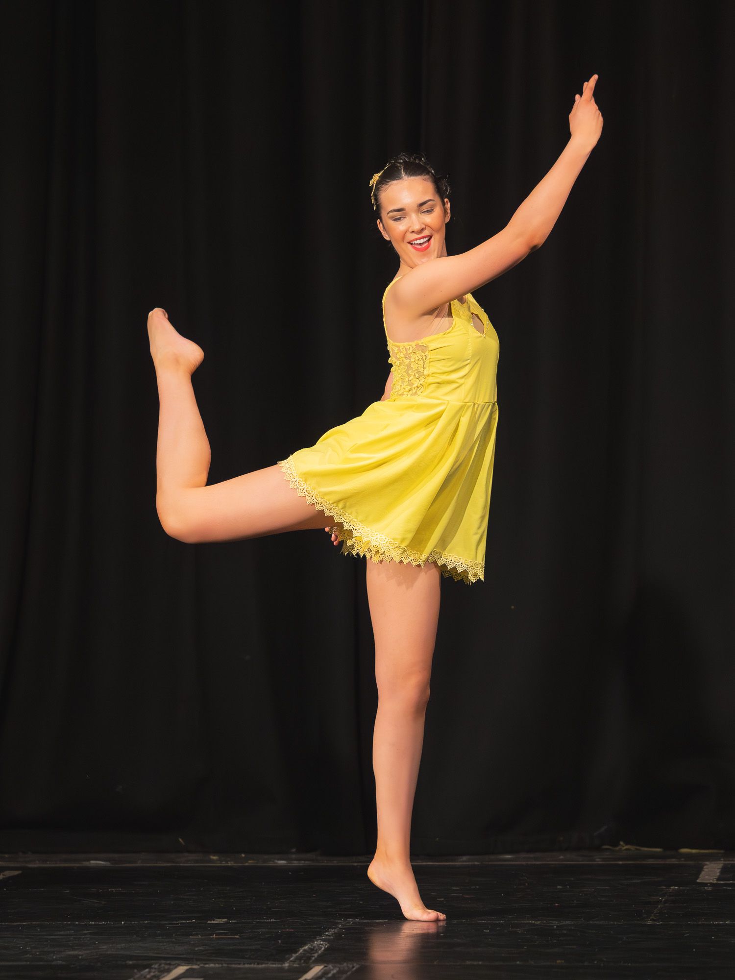 Young dancer in yellow dress performs elegant arabesque on stage against dark curtain backdrop.