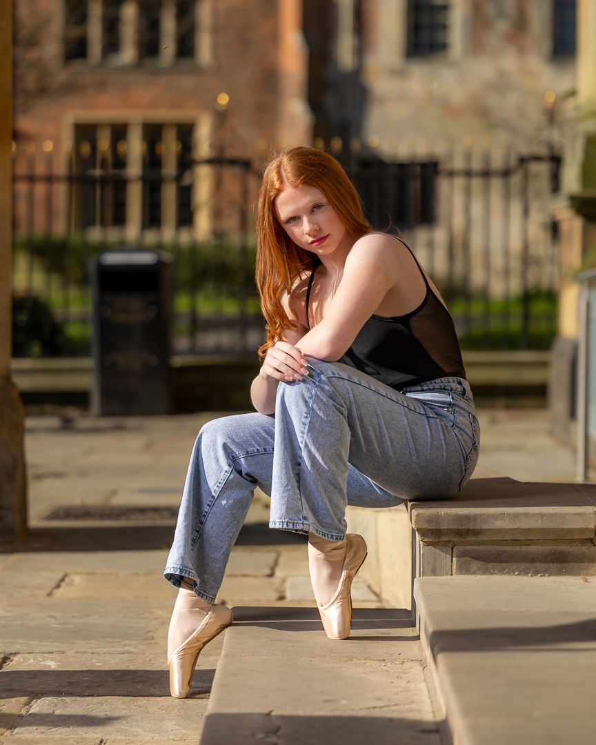 Ballet dancer in casual denim sits thoughtfully on stone steps near historic building.