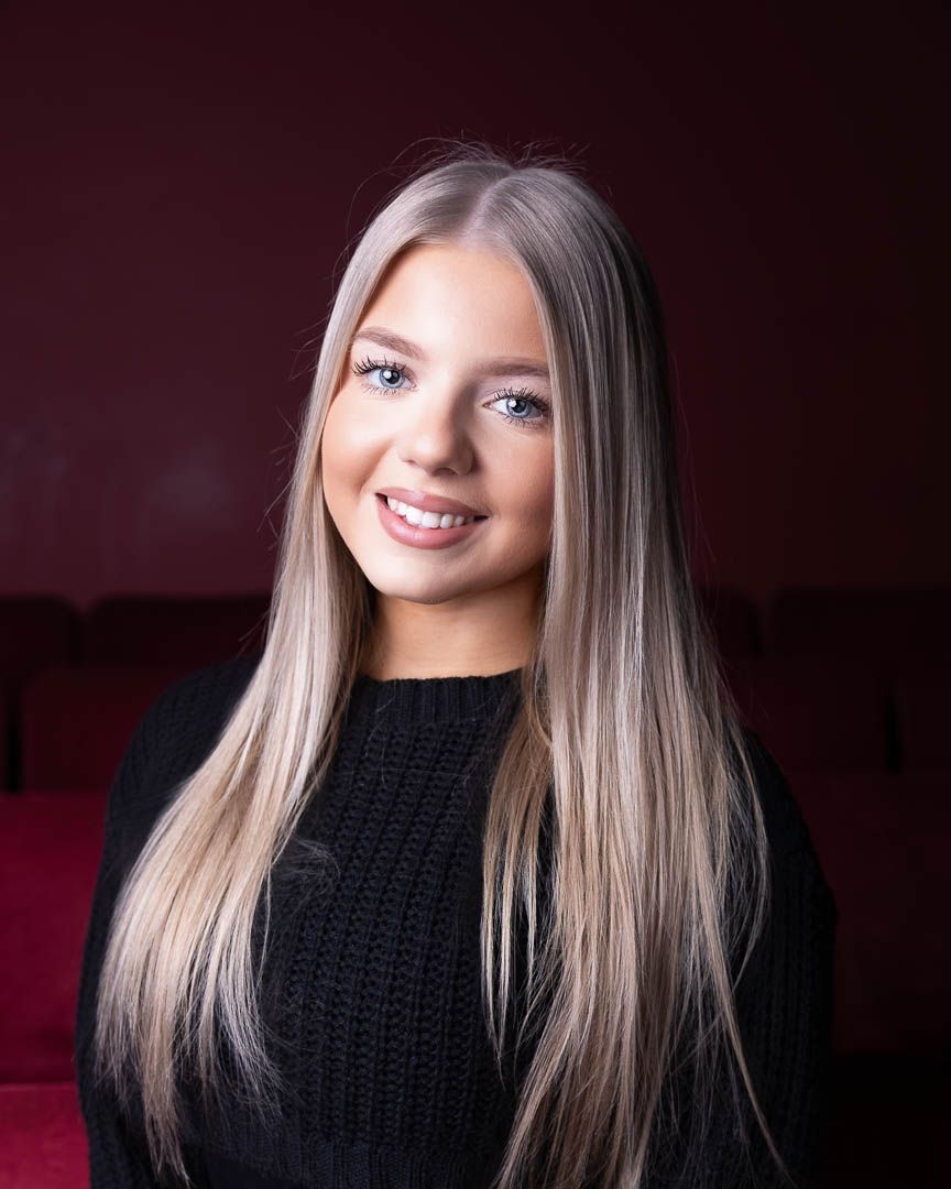 Young woman with long blonde hair and blue eyes smiling, wearing a black sweater against a dark red background.