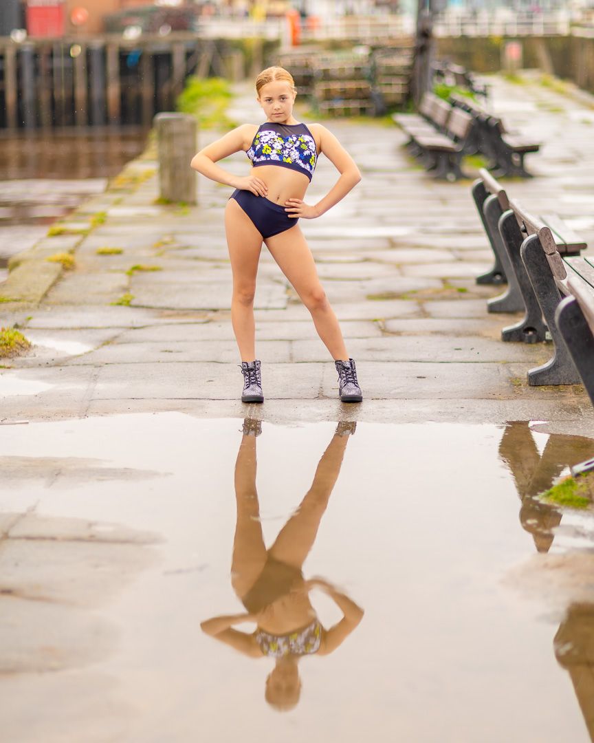 Dancer poses confidently in colorful crop top and shorts, reflected in a puddle on a wet urban waterfront.