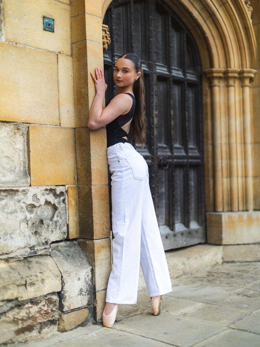 Young woman in white wide-leg pants and black top posing by a stone archway door.