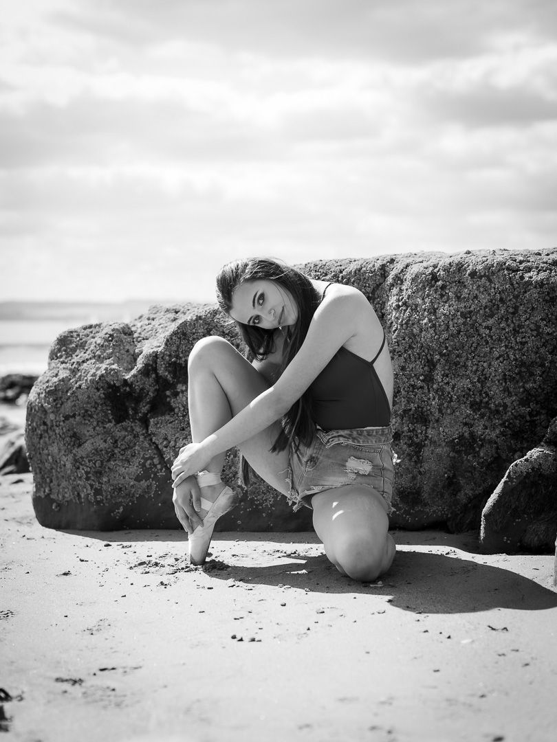 Young woman posing gracefully on sandy beach beside large rocks in black and white.