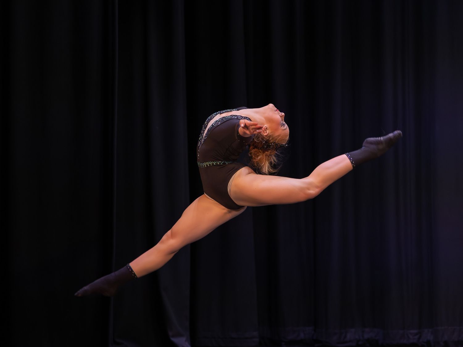 Gymnast performing a stunning split leap in a dark leotard against a black curtain backdrop on stage.