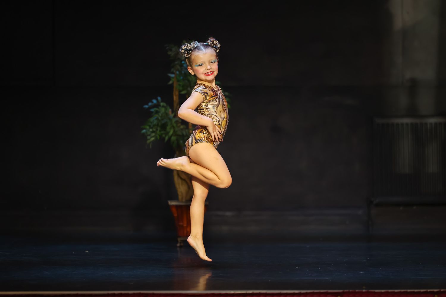 Young girl in gold costume and flower crown performs a ballet pose on stage under spotlight.
