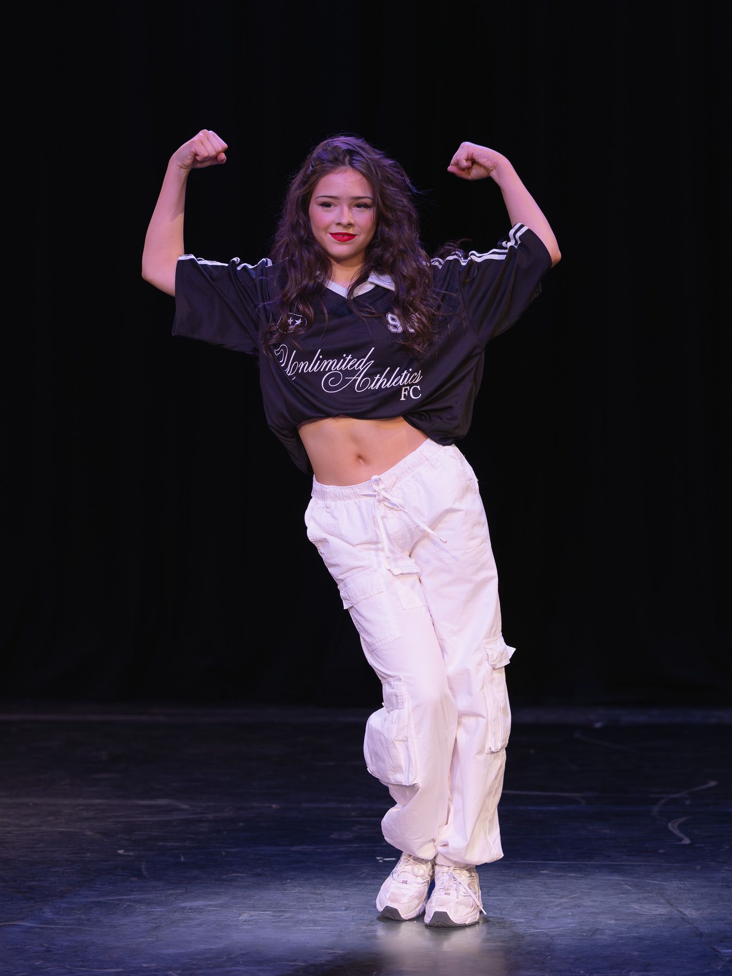 Young dancer on stage flexing biceps in crop top jersey and white baggy pants performing hip-hop routine.