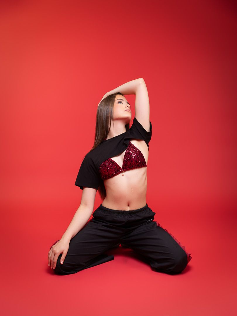 Young woman in black outfit and red sequin bralette poses gracefully on knees against vibrant red background.
