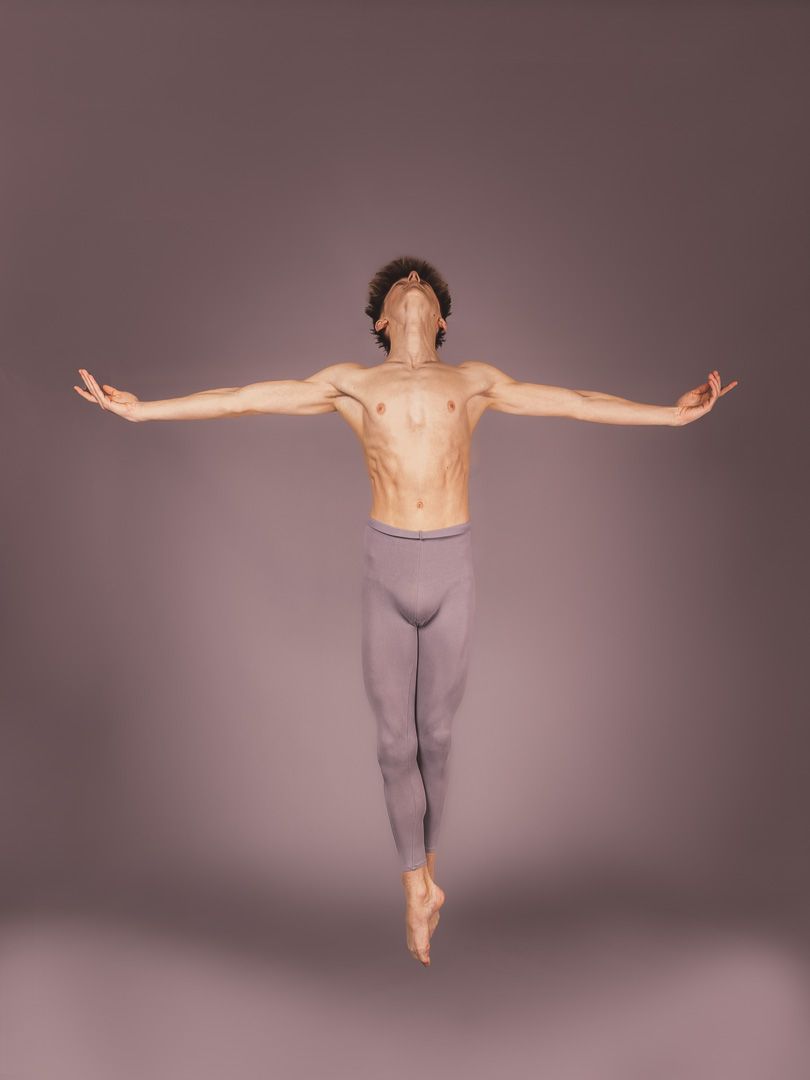 A male ballet dancer leaps gracefully with arms outstretched, shirtless in gray tights, against a soft mauve studio background.
