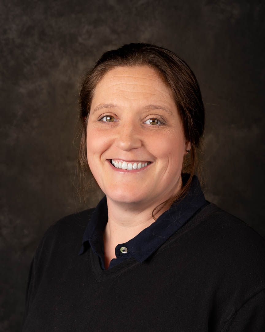 Professional headshot of a smiling woman with brown hair wearing a dark navy top against a gray background.